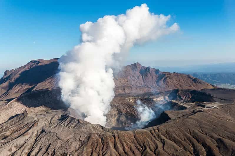 Veduta aerea di un vulcano attivo con un'ampia colonna di fumo bianco che si alza dal cratere sotto un cielo azzurro.