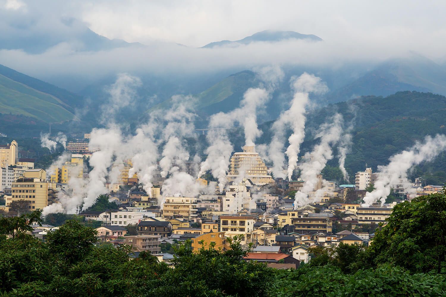 Fumi di vapore salgono dagli edifici di una città adagiata su uno sfondo di montagne verdi e nebbiose sotto un cielo nuvoloso.