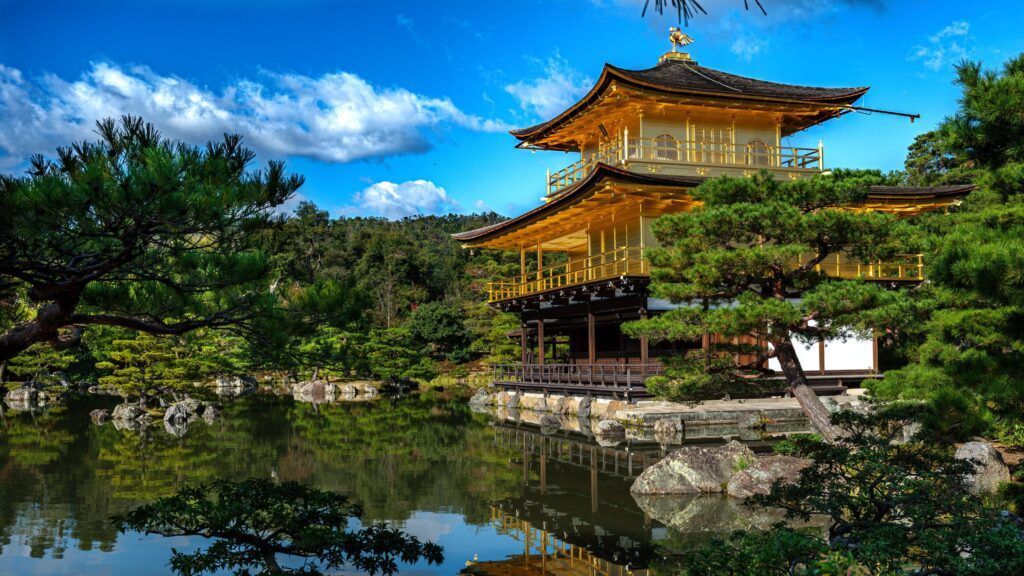 Un tempio dorato a più livelli si riflette in un lago calmo, circondato da alberi verdi sotto un cielo blu.