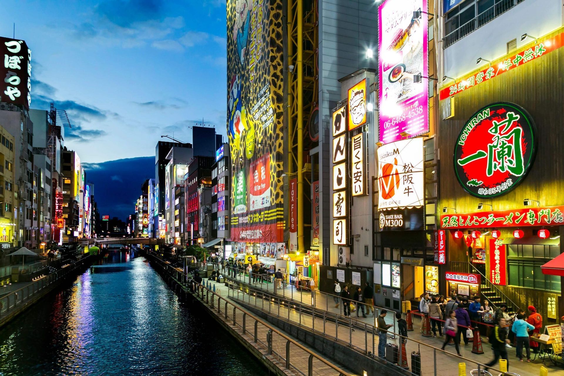 Un canale urbano al tramonto, con persone che passeggiano su un lungofiume fiancheggiato da edifici illuminati da insegne al neon.