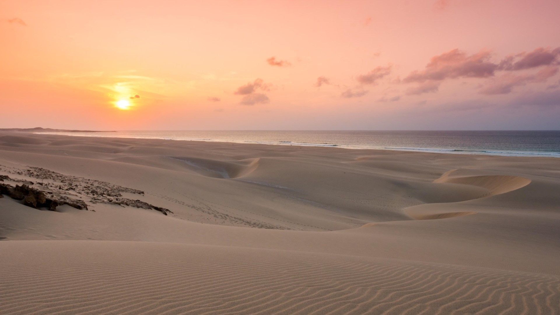 El sol se pone sobre el océano, proyectando un resplandor rosa y naranja sobre un vasto paisaje de dunas de arena.
