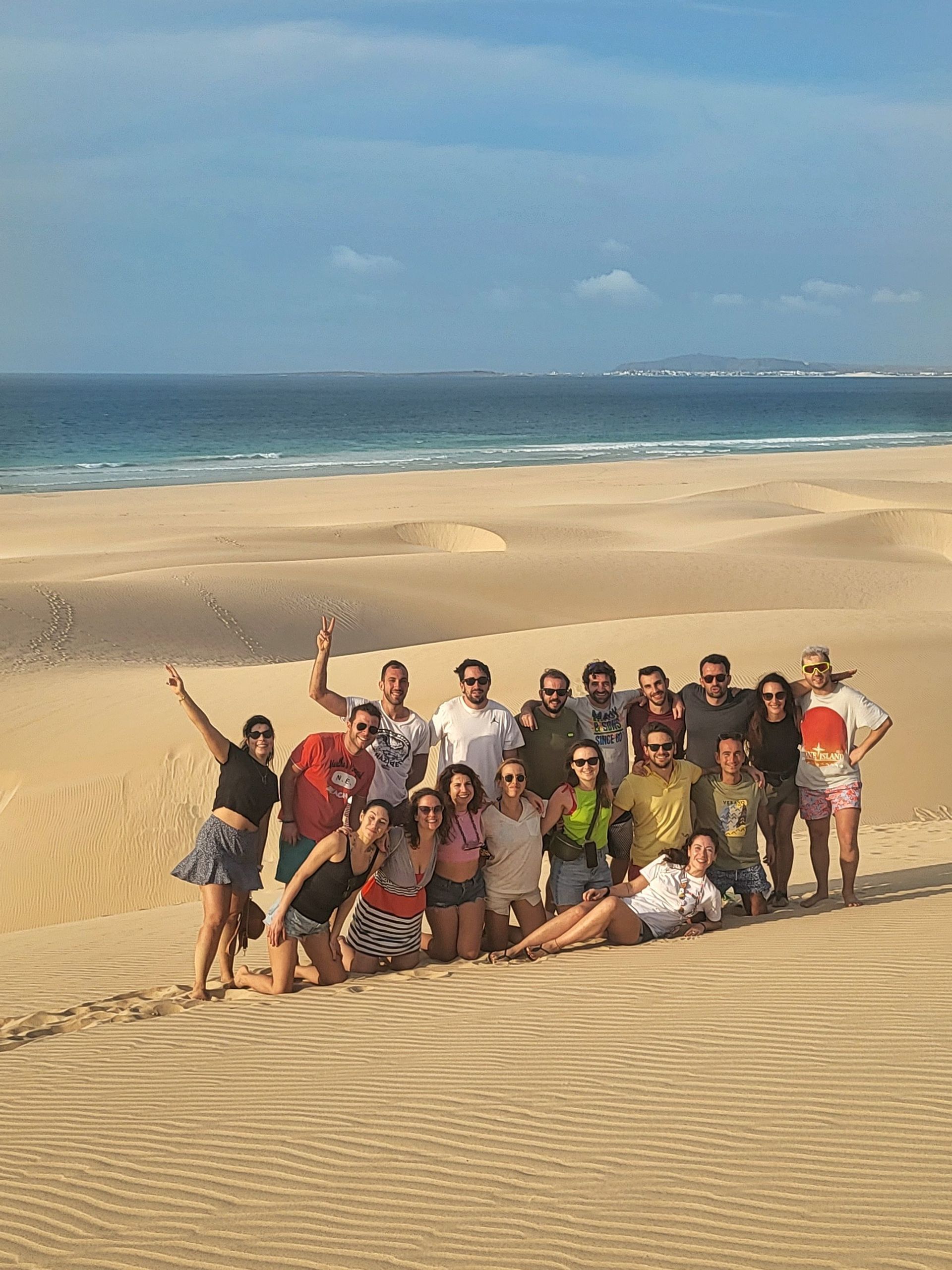Un viaje en grupo de WeRoad posando para una foto en grandes dunas de arena con el océano y una costa distante al fondo.