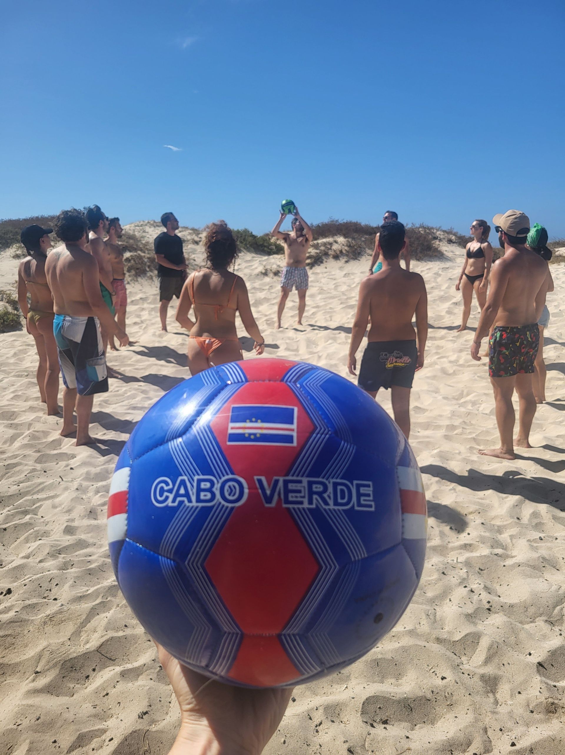 Una mano sostiene un balón de fútbol de Cabo Verde en primer plano, con un viaje grupal de WeRoad jugando juntos en una playa de arena.