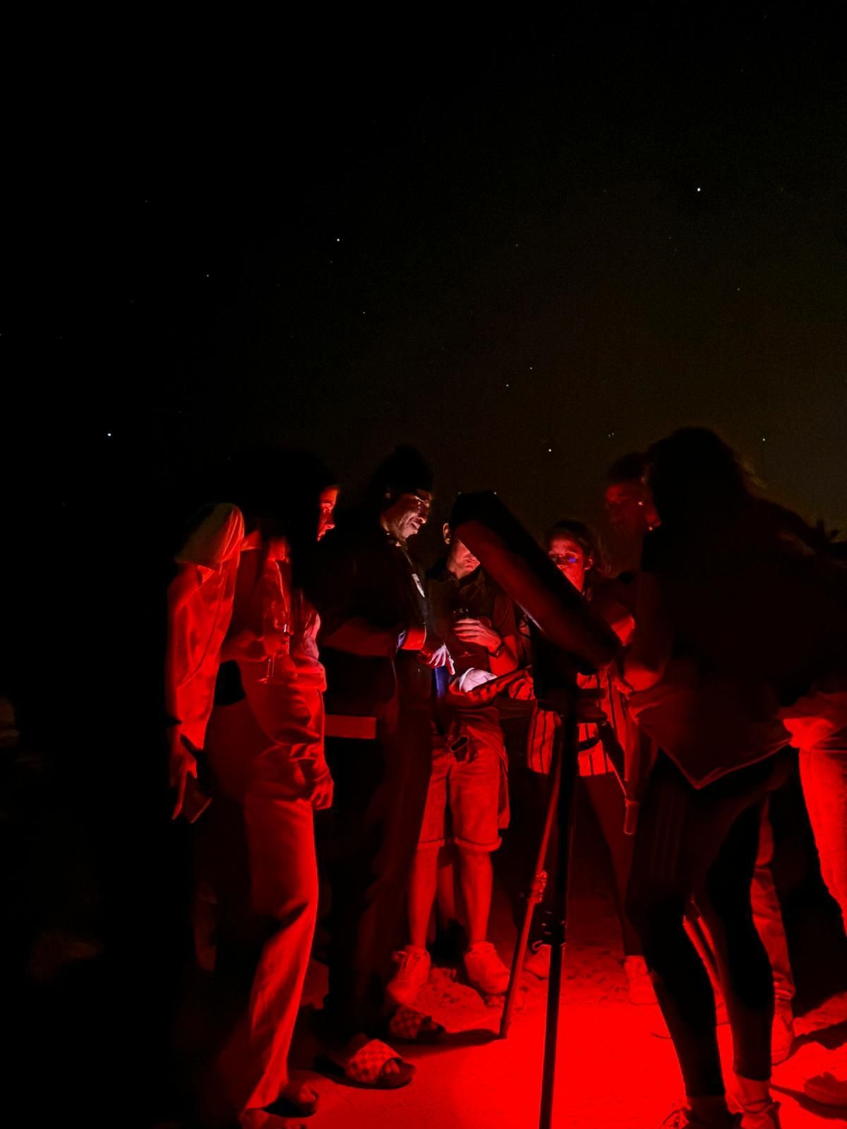Un viaje en grupo de WeRoad se reúne alrededor de un telescopio para observar las estrellas por la noche, iluminado por una luz roja.