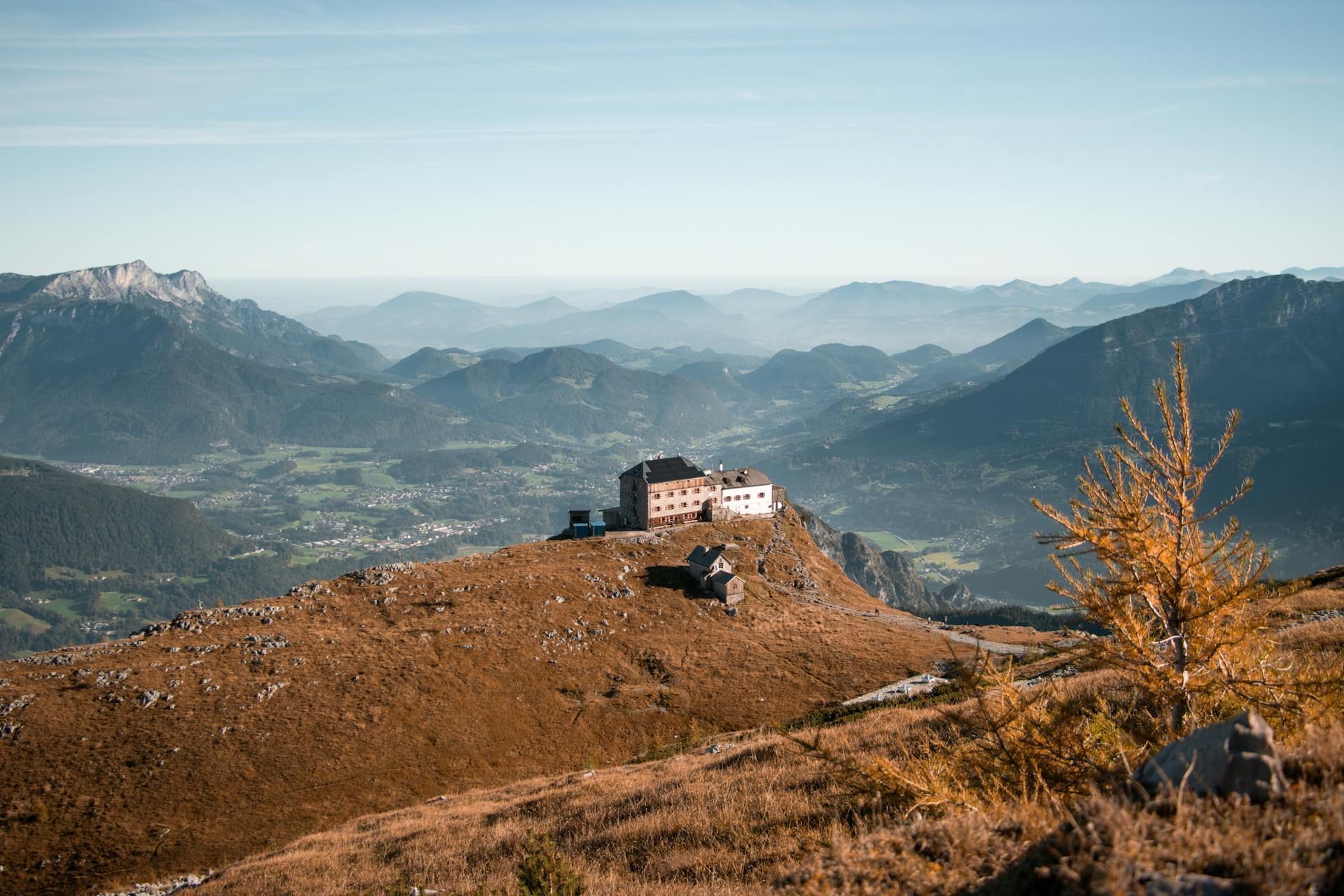 Ein großes Gebäude auf einem Berggipfel überblickt ein weites Tal und Schichten ferner Berge unter einem dunstigen blauen Himmel.