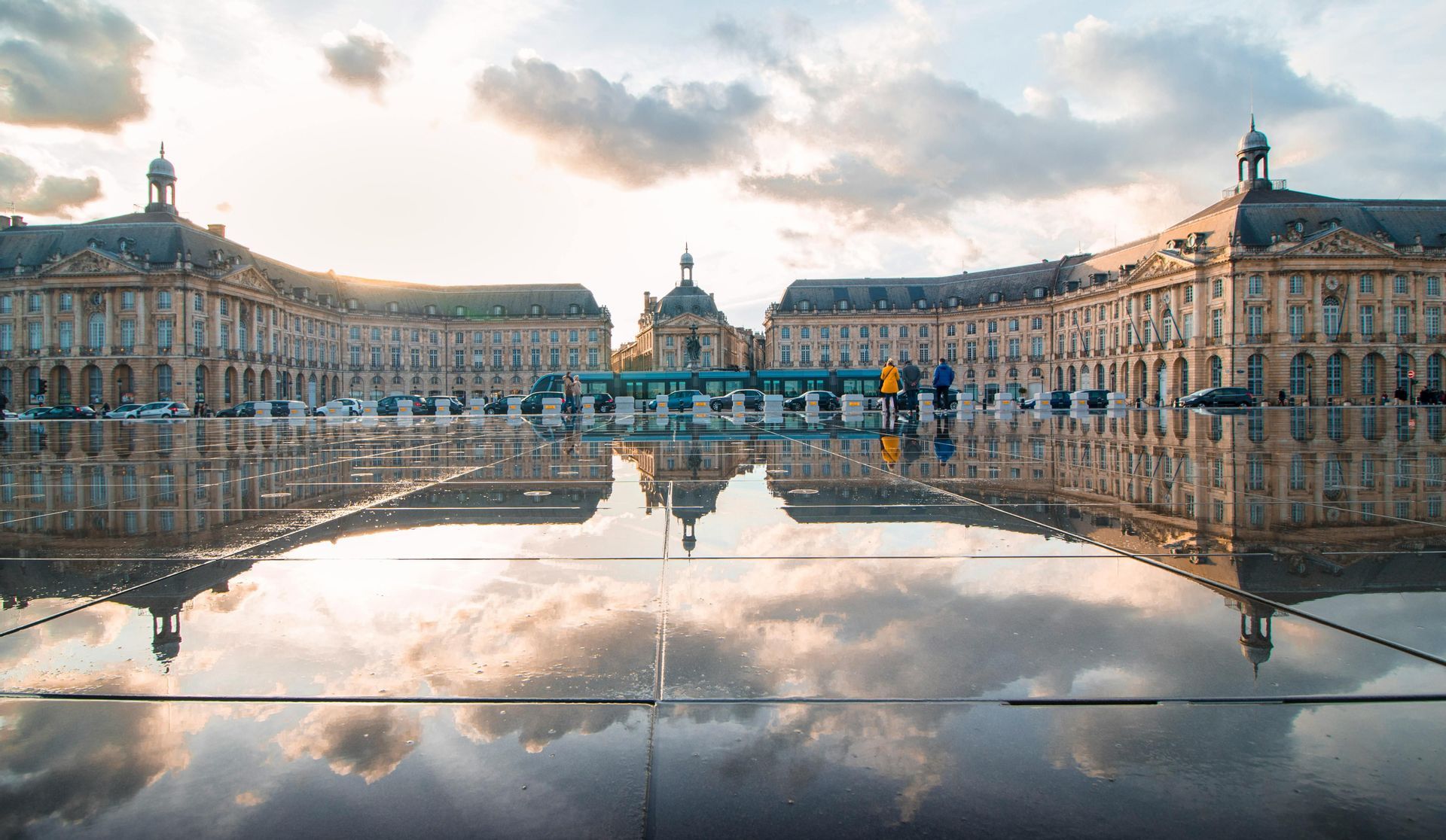 Un gran complejo de edificios clásicos y un cielo nublado se reflejan perfectamente en una plaza mojada al atardecer.