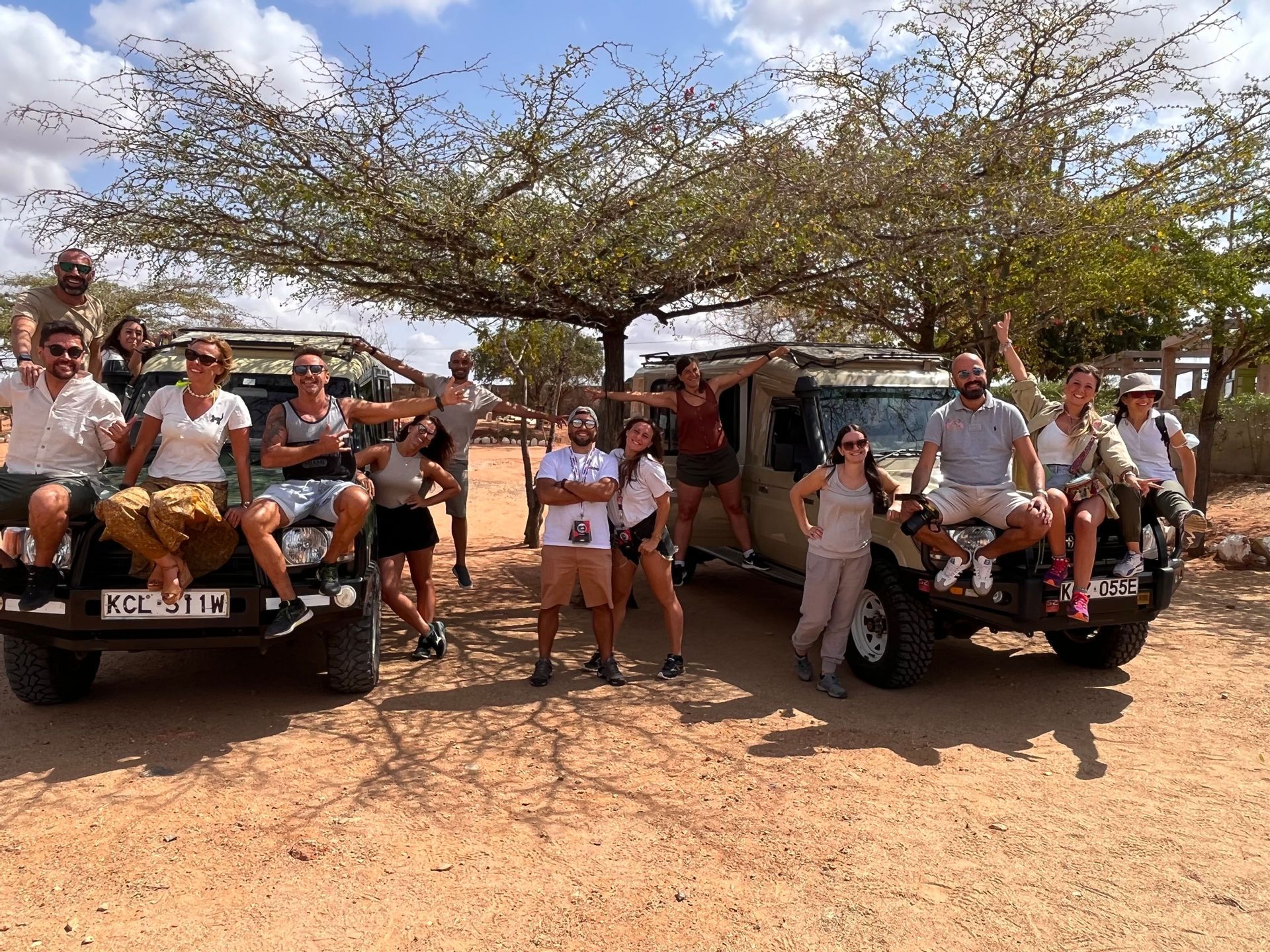 Un gruppo WeRoad in posa per una foto con due jeep safari sotto un grande albero in un paesaggio polveroso.