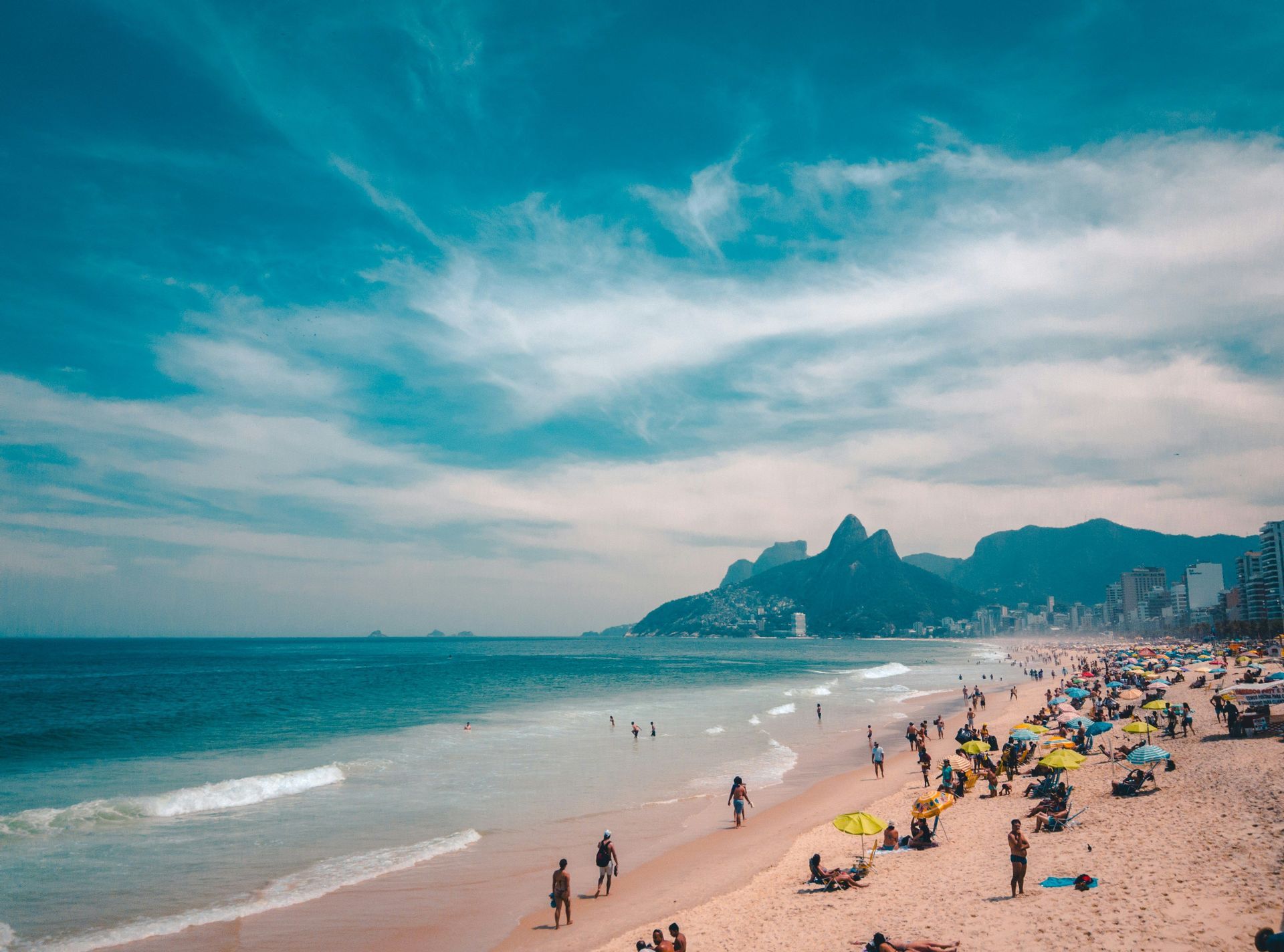 Una spiaggia sabbiosa affollata con persone che prendono il sole e nuotano, con una città e montagne sullo sfondo sotto un cielo azzurro brillante.