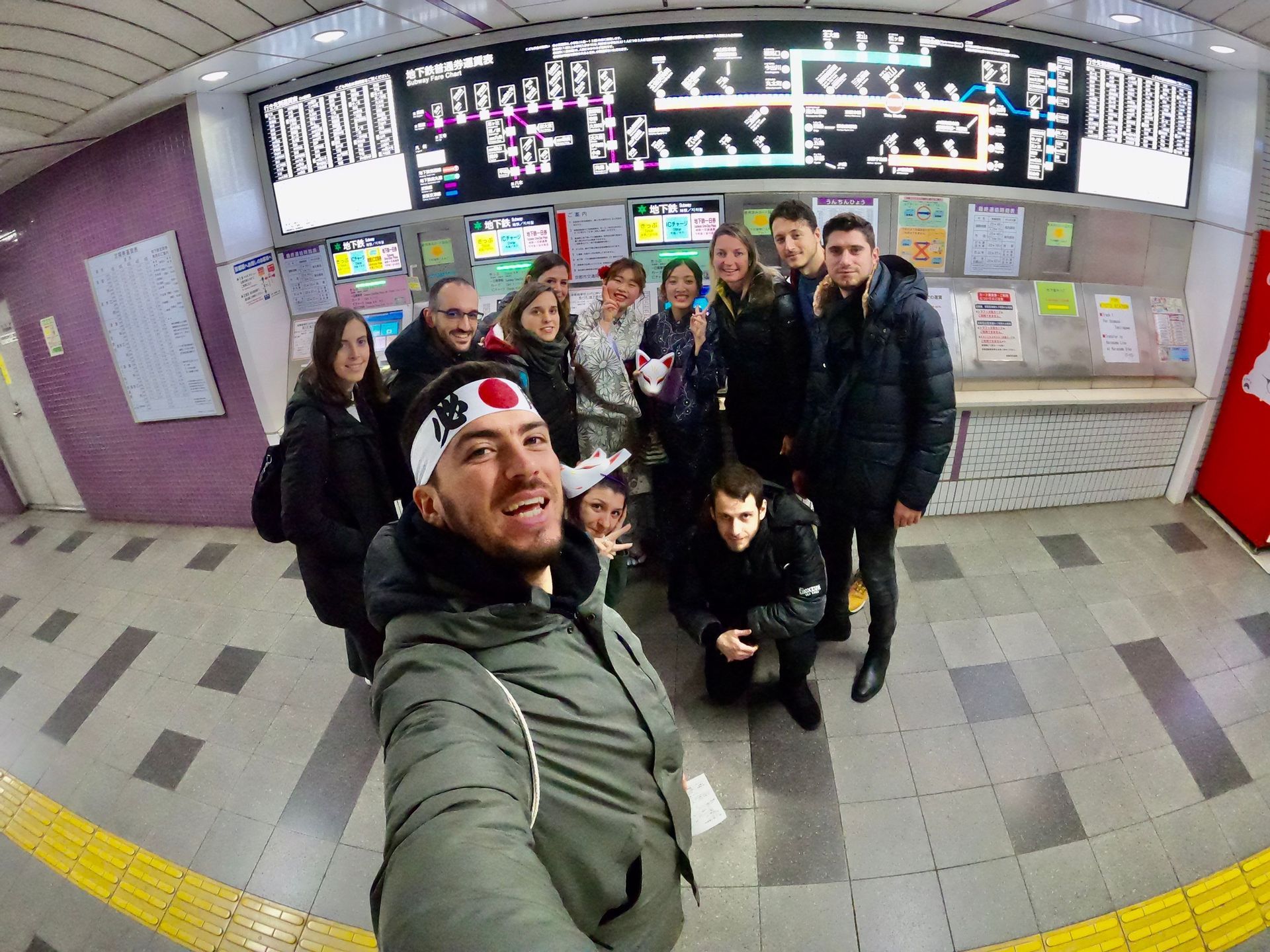 Un groupe WeRoad en voyage fait un selfie de groupe dans une station de métro, devant une grande carte électronique.