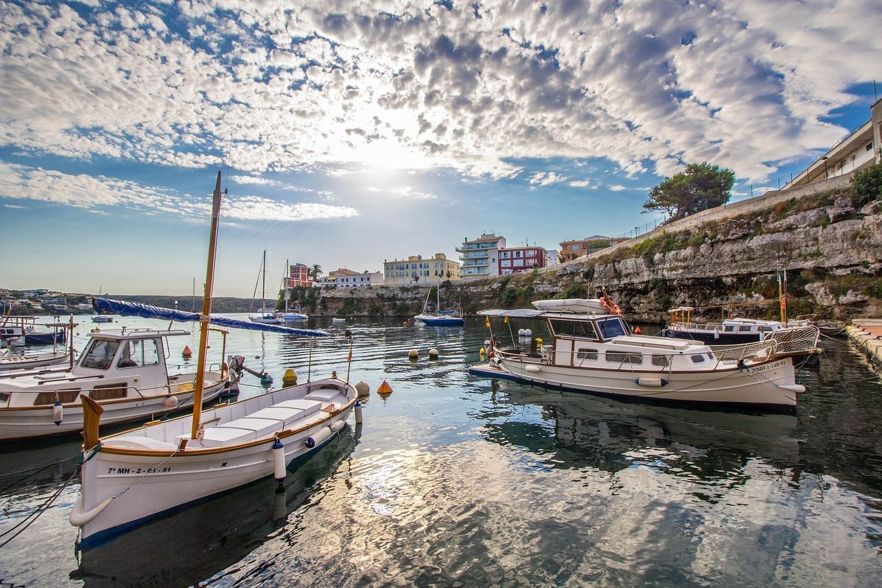 Plusieurs bateaux sont amarrés dans un port calme, près d'une falaise avec des bâtiments, sous un ciel partiellement nuageux.