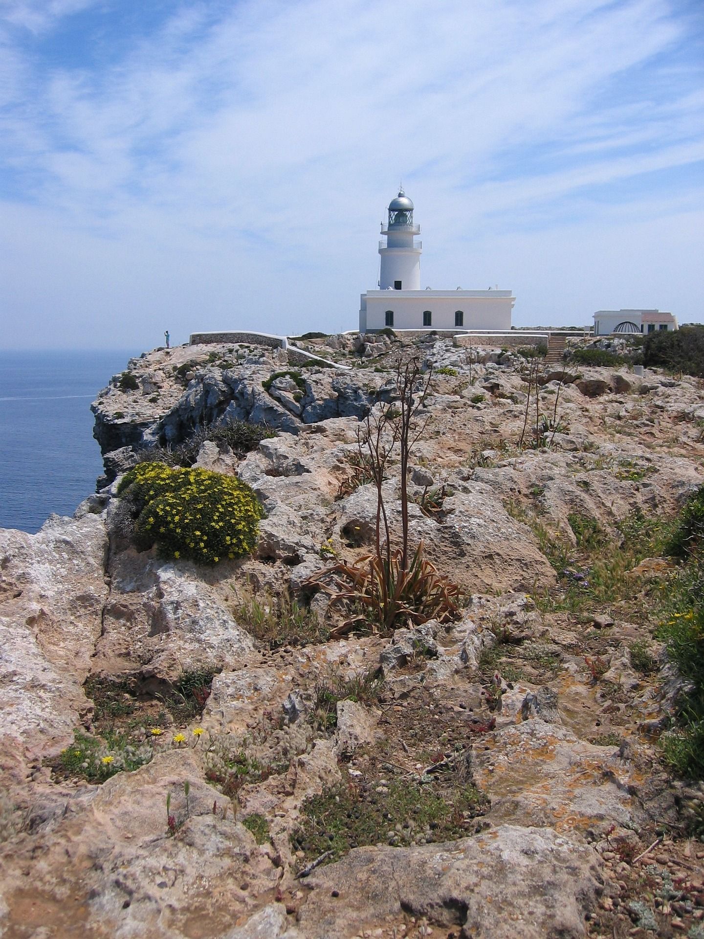 Ein weißer Leuchtturm thront auf einer felsigen Klippe mit Blick auf das blaue Meer unter einem teils bewölkten Himmel.