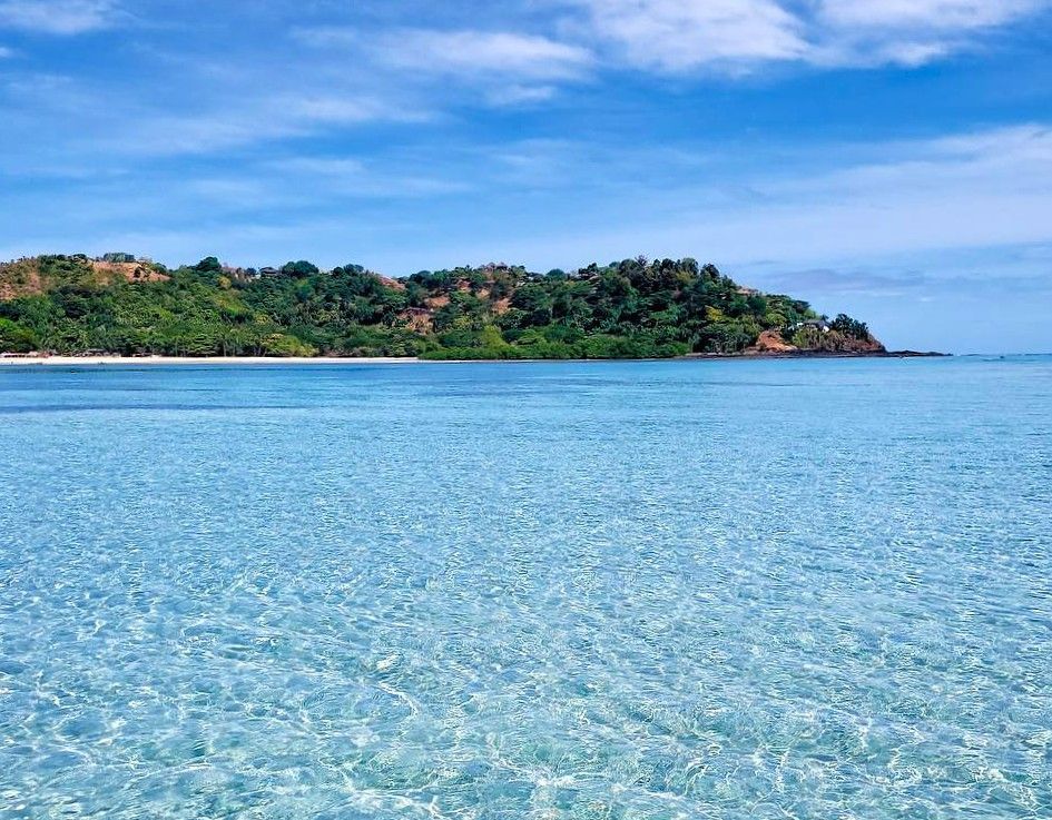 Acqua cristallina color turchese luccica davanti a un'isola verde lussureggiante con una piccola spiaggia di sabbia bianca sotto un cielo blu.