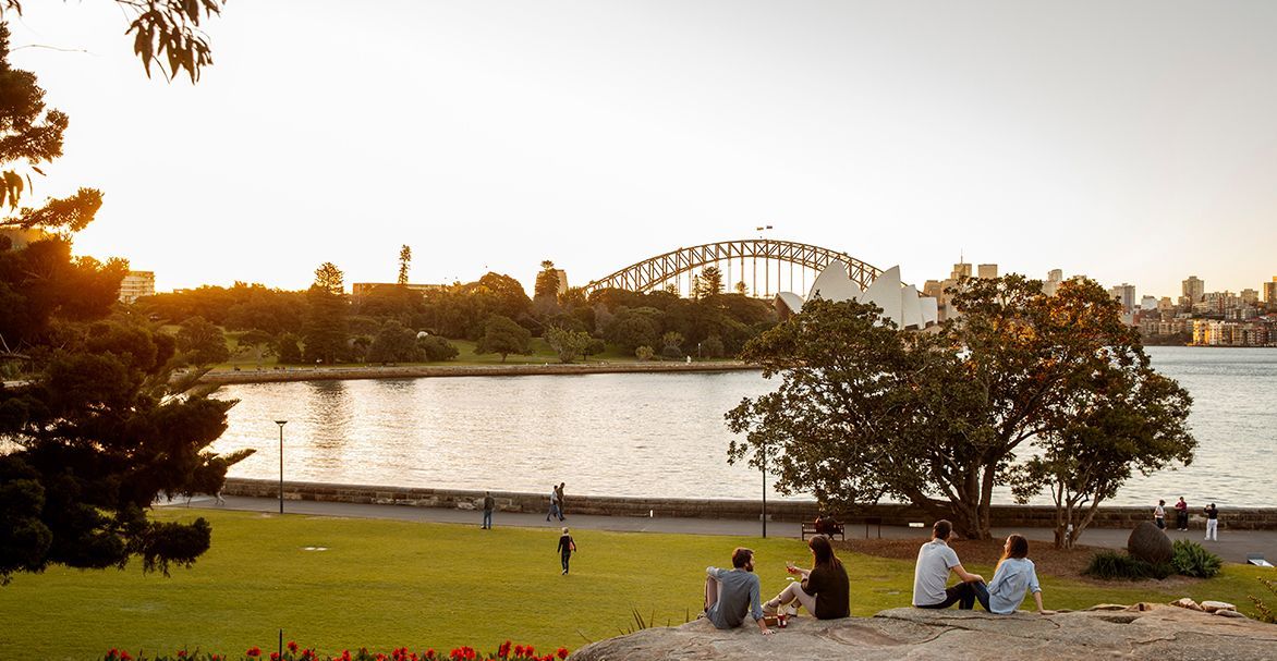 Un grupo de WeRoad de viaje relajándose en una roca en un parque, disfrutando de la puesta de sol con vistas al horizonte de la ciudad y un gran puente sobre el agua.