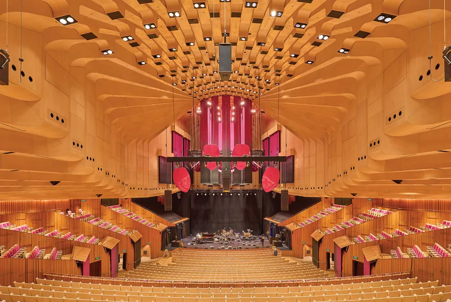 El interior de una sala de conciertos vacía con paredes y techo de madera curvada, asientos escalonados y un escenario con instrumentos musicales.