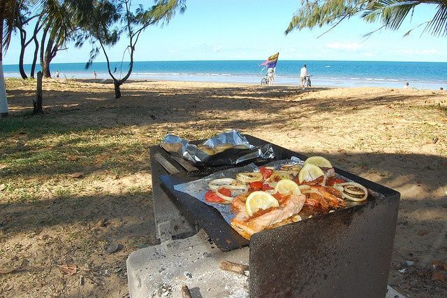 Salmón a la parrilla con verduras y rodajas de limón cocinándose en una barbacoa en una playa de arena junto al océano.