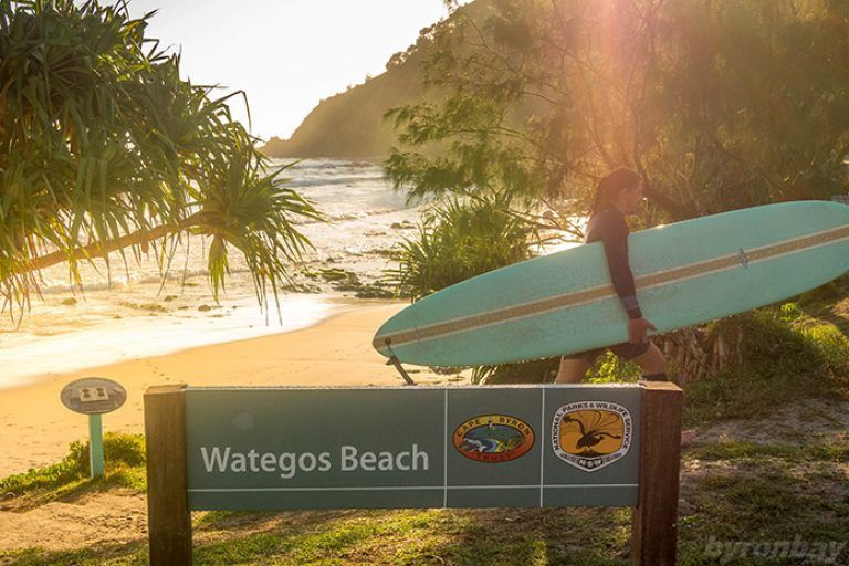 Un surfista con traje de neopreno lleva una tabla de longboard pasando un cartel de la playa de Wategos, caminando hacia el océano al amanecer.