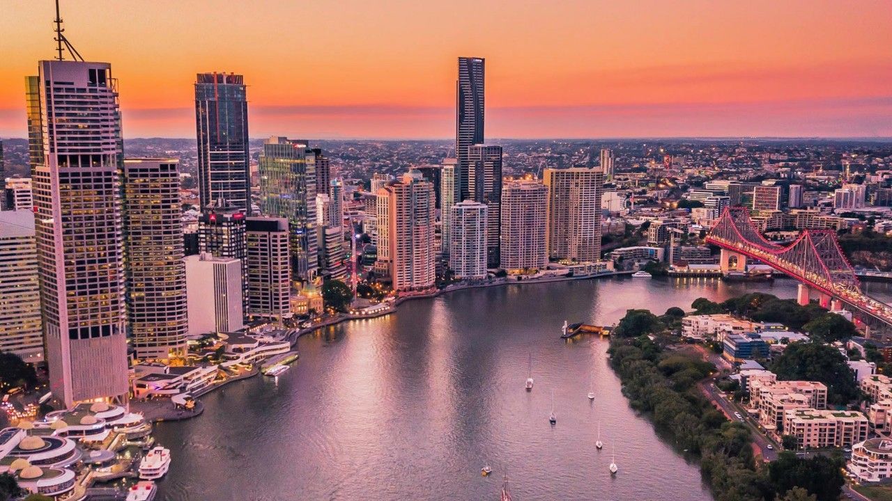 Una vista aérea de un paisaje urbano moderno con rascacielos a lo largo de un río serpenteante, un puente iluminado y veleros bajo un cielo al atardecer.