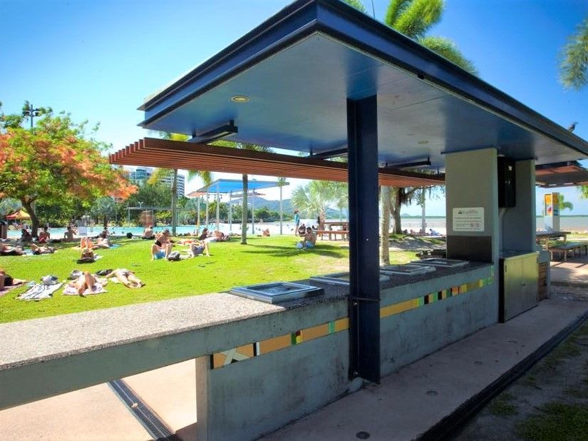 Un refugio público con barbacoas ofrece vistas a un parque con césped donde la gente se tumba al sol y se baña en una laguna en un día soleado.