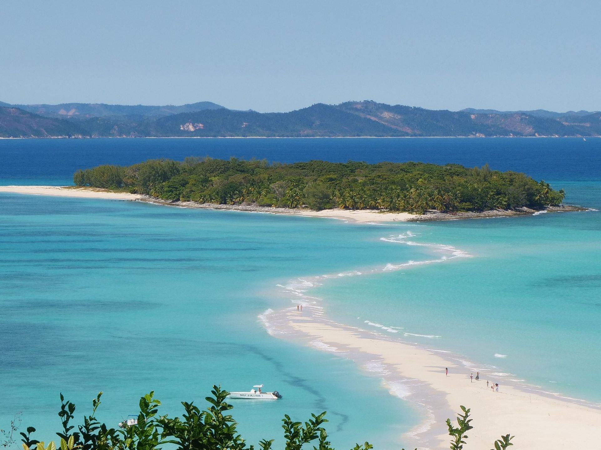 Veduta aerea di una sinuosa lingua di sabbia bianca che porta a un'isola tropicale lussureggiante, con alcune persone che vi camminano e una barca nel mare turchese.