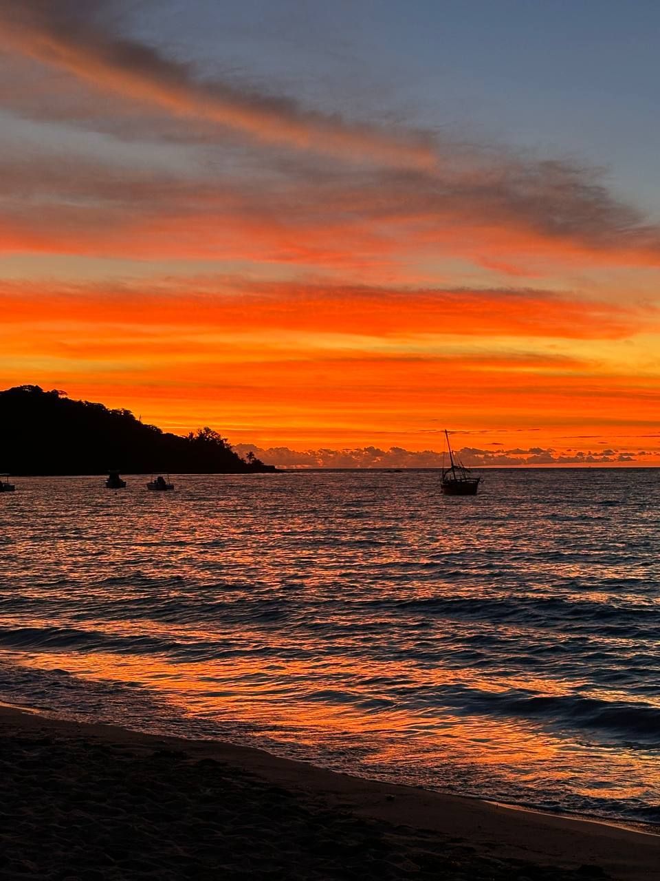 L'oceano riflette un tramonto vibrante arancione e rosso, mentre diverse barche sono stagliate contro una costa scura.