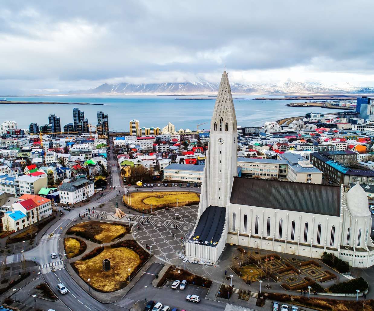 Una vista aerea di una grande chiesa moderna che domina una colorata città costiera con montagne innevate in lontananza.