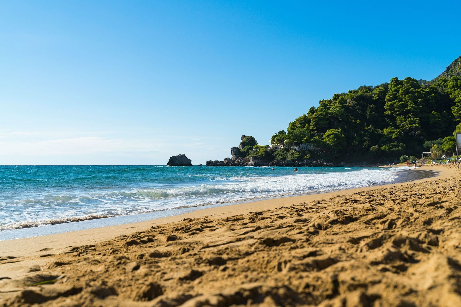 Una vista dal basso di una spiaggia di sabbia con onde dolci, un mare turchese e una costa verde e alberata sotto un cielo azzurro e sereno.