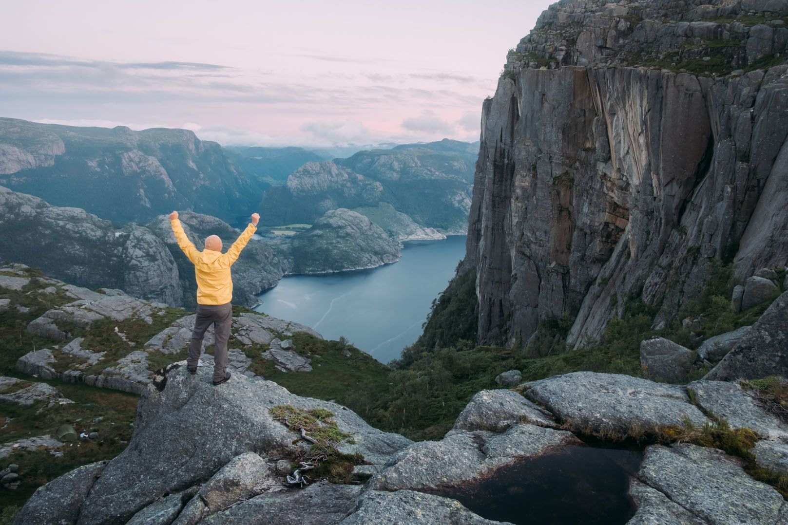 Une personne en veste jaune se tient sur un promontoire rocheux, les bras levés, surplombant un fjord et un paysage montagneux.