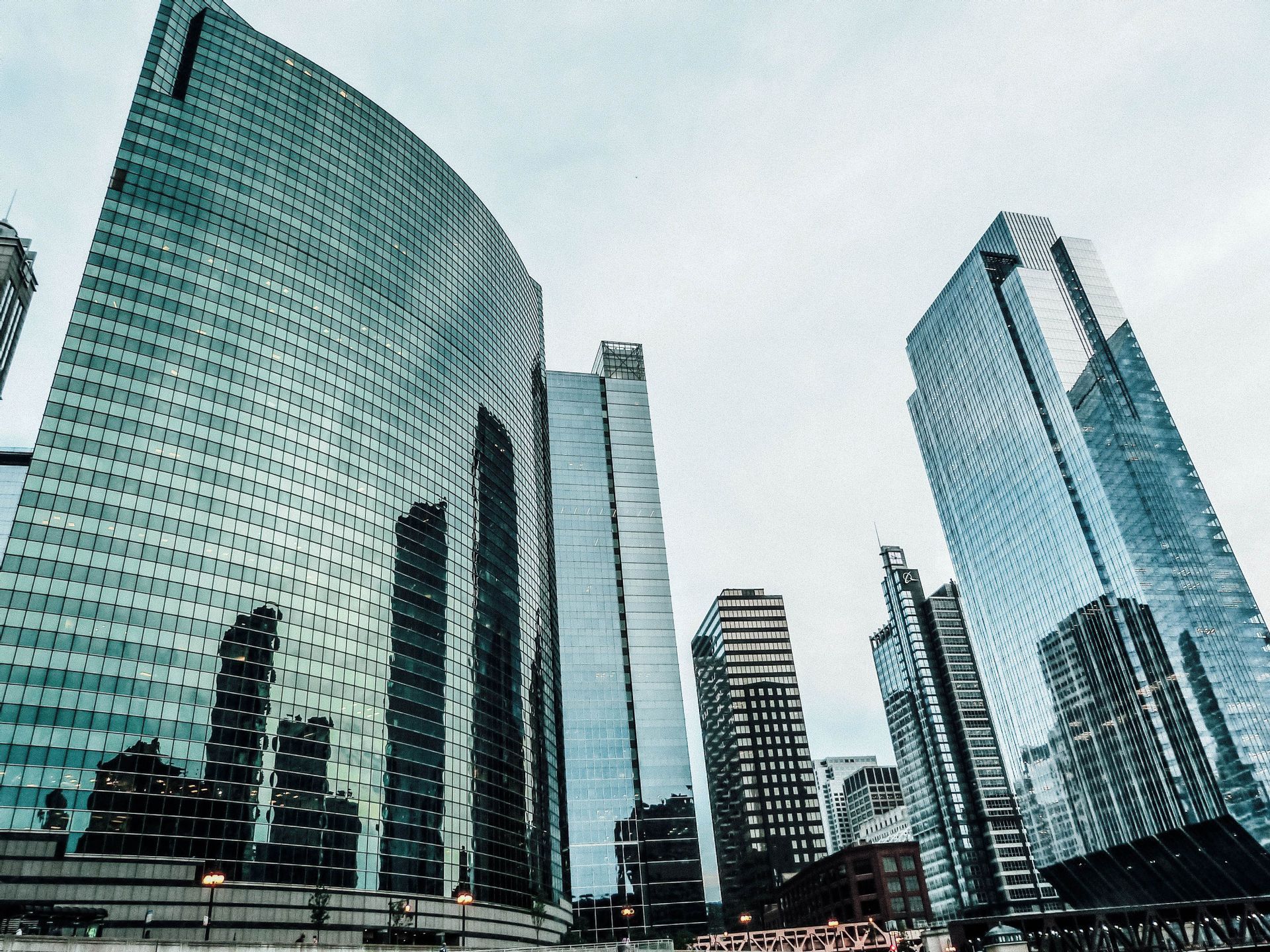Blick aus der Froschperspektive auf aufragende Glaswolkenkratzer mit gekrümmter Fassade, die andere Gebäude vor bedecktem Himmel spiegelt.