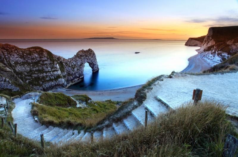 Una scala in pietra scende da una scogliera erbosa verso una spiaggia con un grande arco di roccia nell'oceano al tramonto.