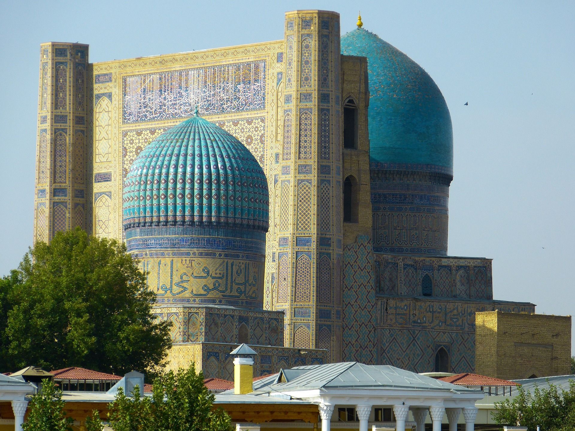 Un edificio sontuoso con grandi cupole turchesi e minareti decorati con intricati mosaici, sullo sfondo di un cielo limpido.