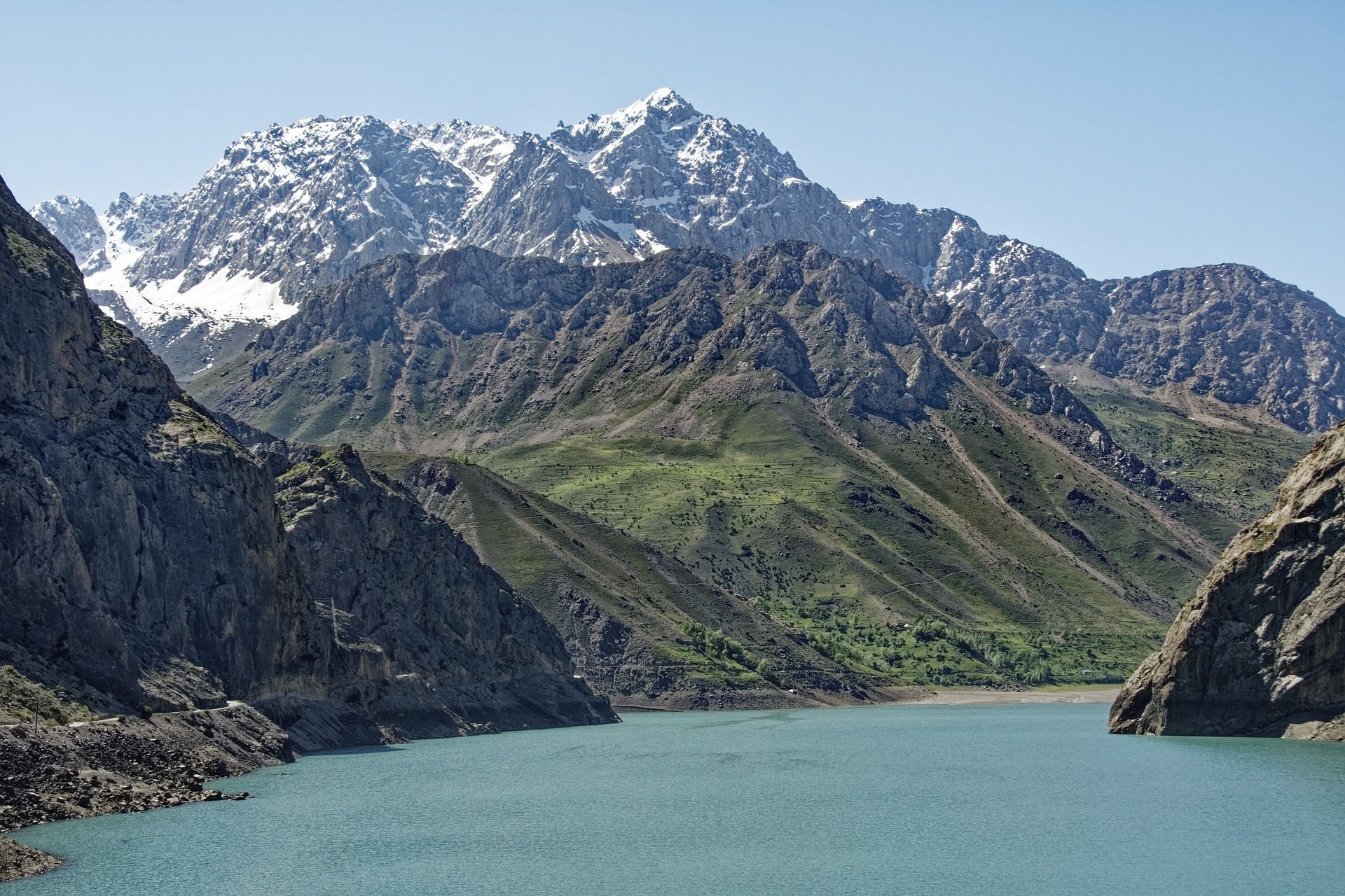 Un lago turchese in una valle circondata da ripidi pendii verdi e montagne rocciose, con cime innevate sullo sfondo.