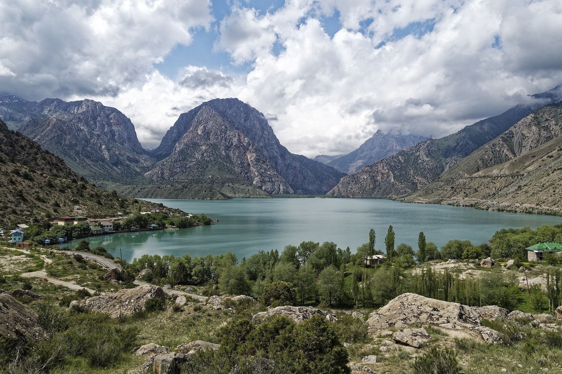 Un lago turchese si trova in una valle tra montagne rocciose, con un piccolo villaggio sulla riva sotto un cielo nuvoloso.
