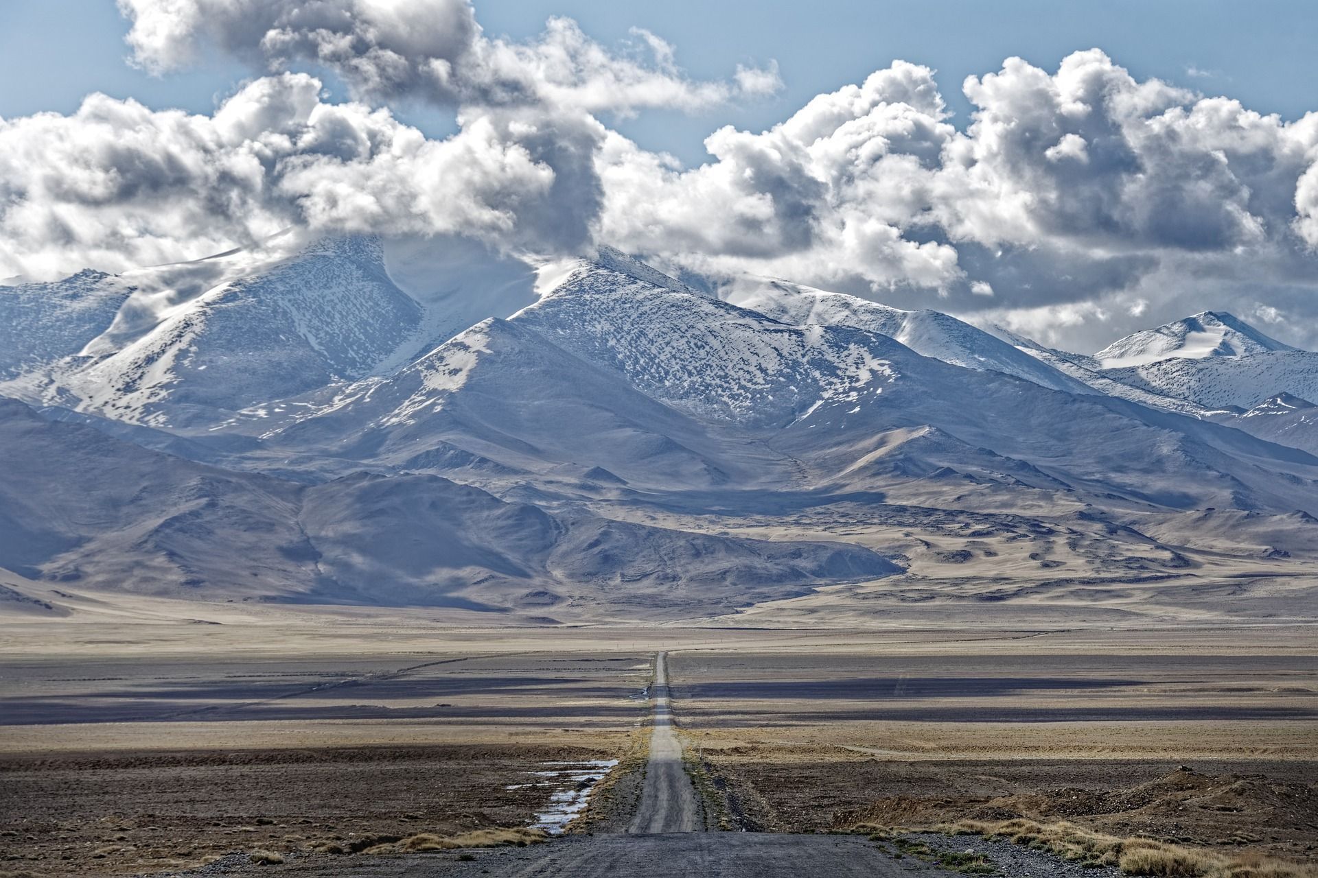 Una lunga e dritta strada sterrata si estende attraverso una vasta pianura arida verso una catena montuosa innevata sotto un cielo nuvoloso.
