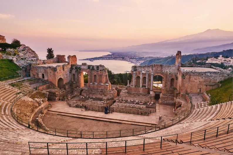Un antico anfiteatro in pietra in rovina, che si affaccia su una costa e una grande montagna in lontananza al tramonto.