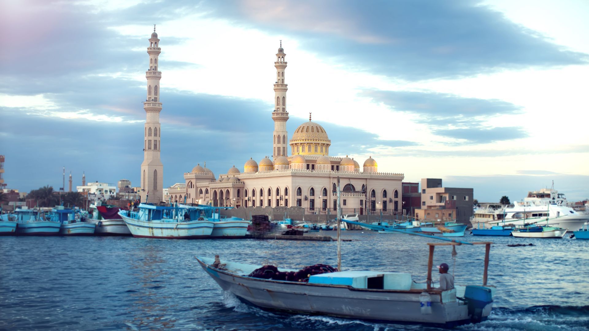 Un motoscafo attraversa un porto con numerose imbarcazioni ormeggiate e, sulla riva, una grande moschea con due minareti e cupole.