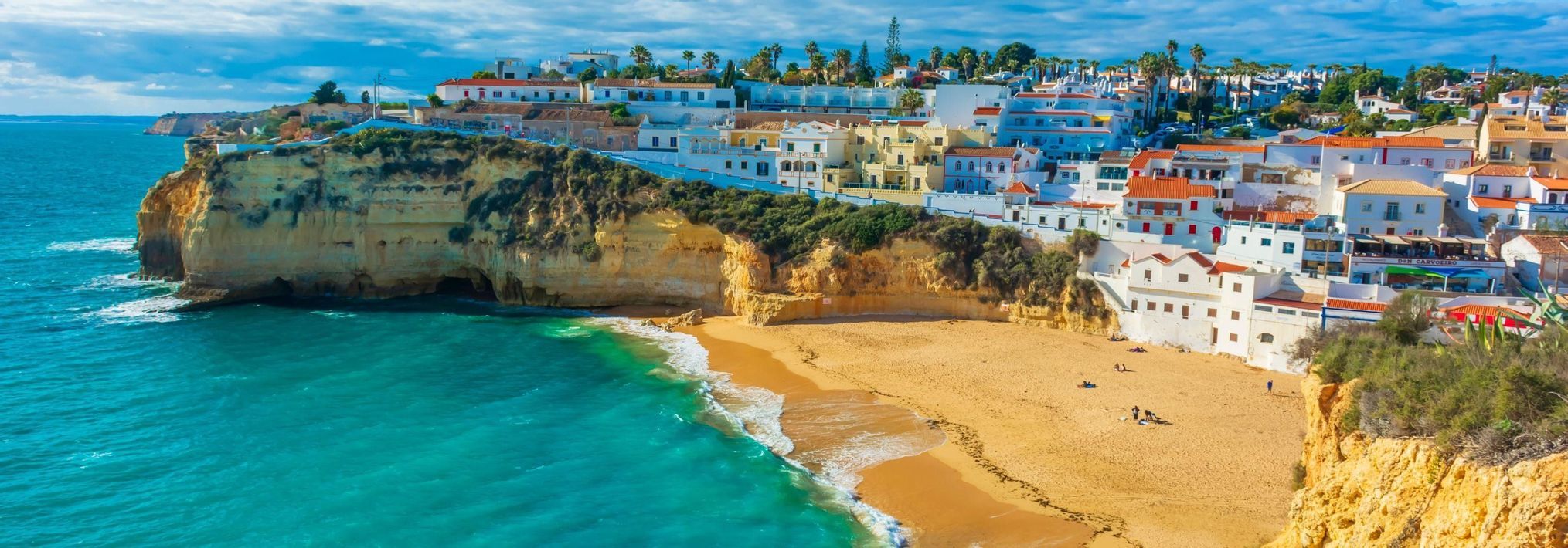 A coastal town with white buildings perched on cliffs above a golden sand beach and a turquoise ocean.