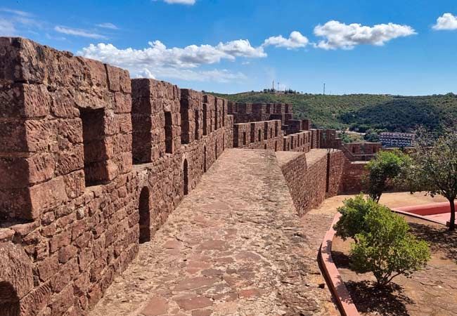 A stone walkway runs along the top of a red brick fortress wall, with a view of green hills under a blue sky.