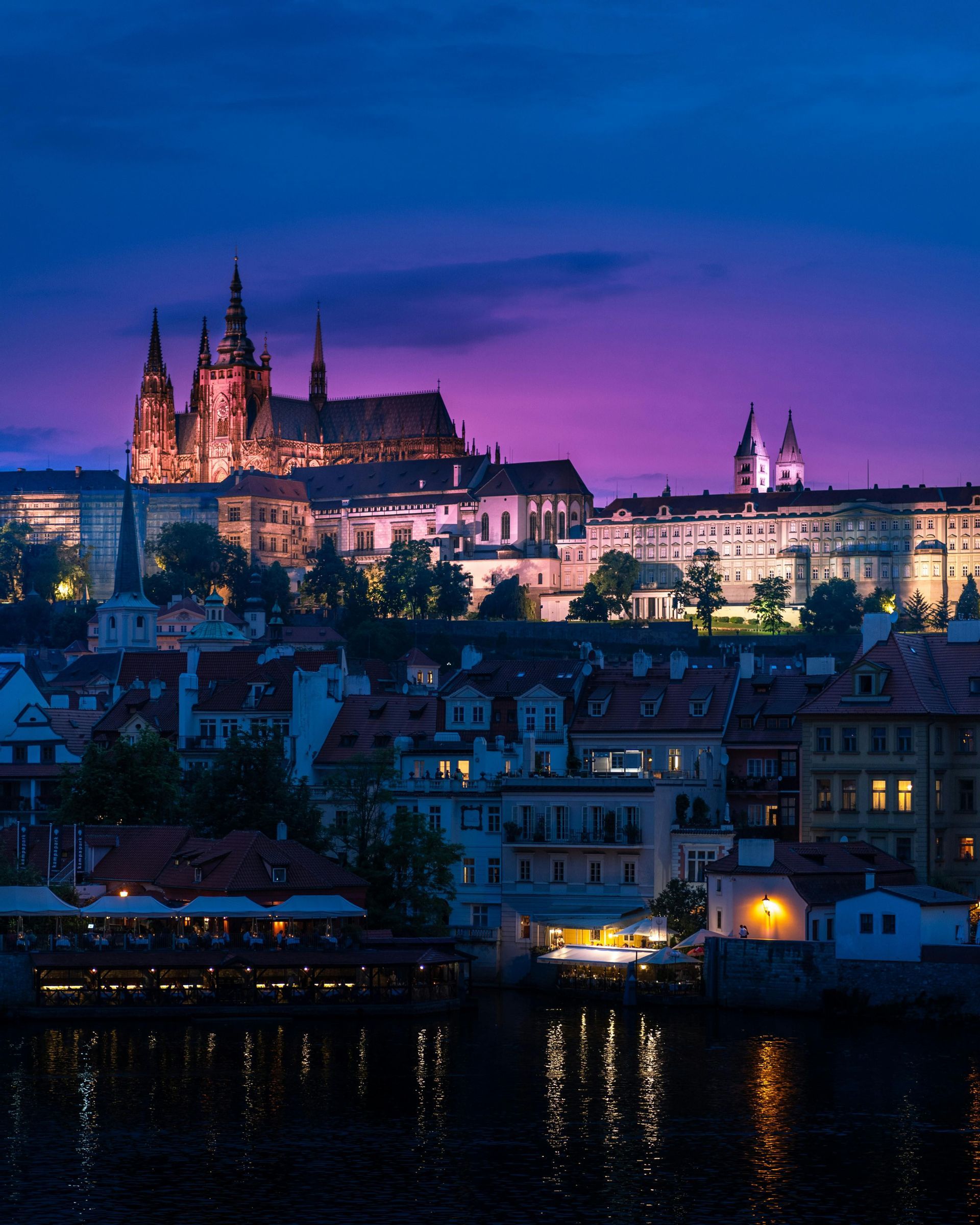 Un castello e una cattedrale illuminati su una collina che domina gli edifici della città e un fiume al crepuscolo sotto un cielo viola