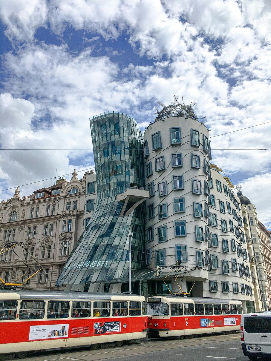 Un moderno edificio decostruttivista con una torre di vetro curva, con due tram rossi e bianchi che passano in primo piano.