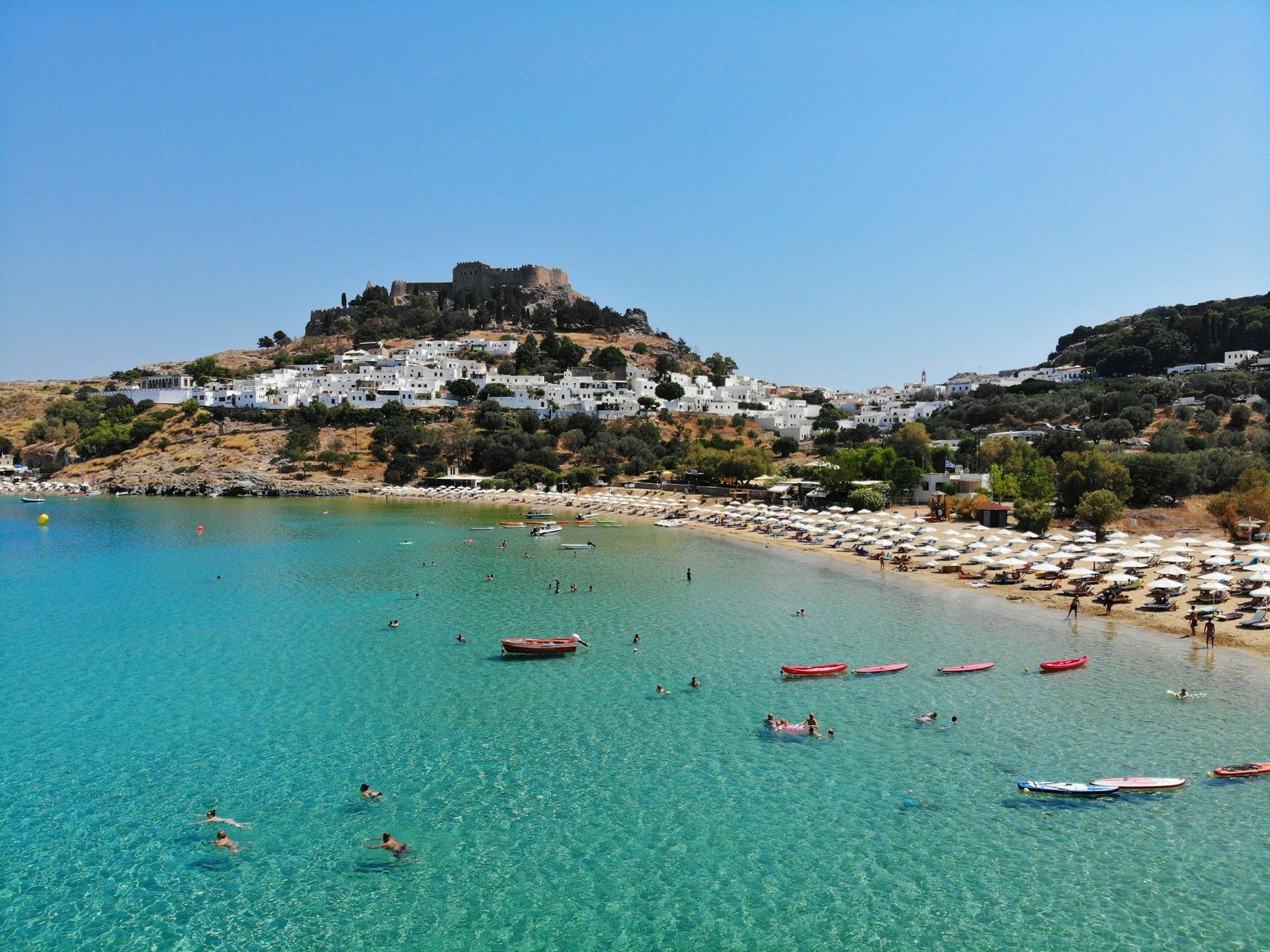 Una vista aerea di una baia con acqua turchese, una spiaggia sabbiosa con ombrelloni bianchi e un villaggio collinare con una fortezza in cima.