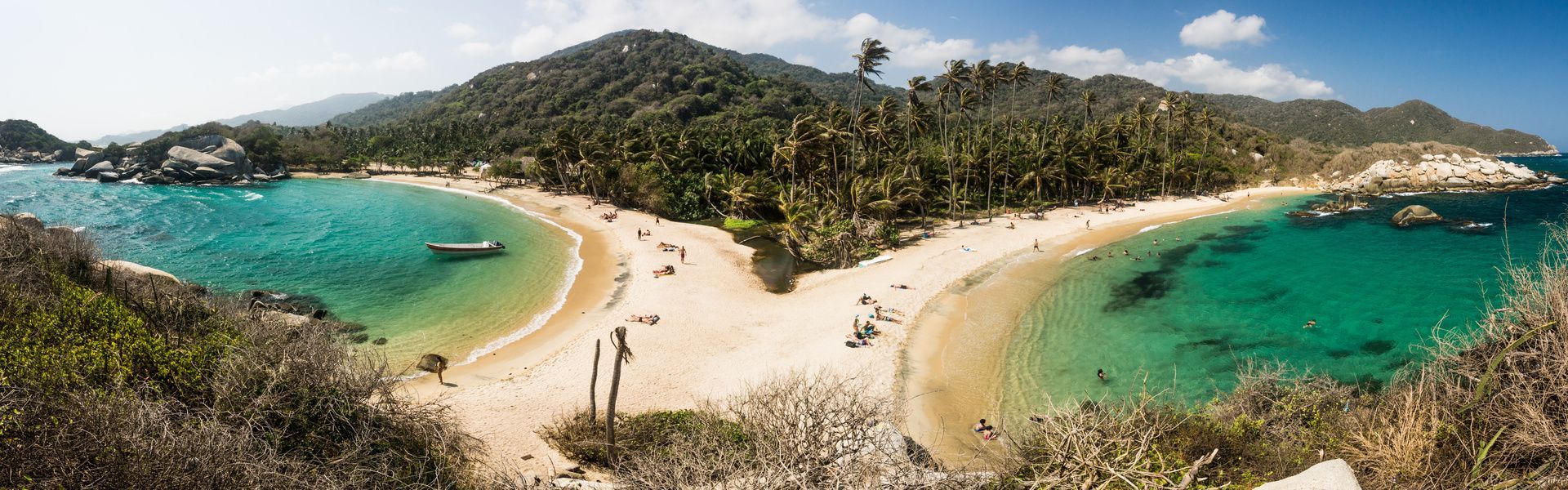 Un uomo siede su una grande roccia che domina due spiagge tropicali con acqua turchese, palme e una lussureggiante collina verde.