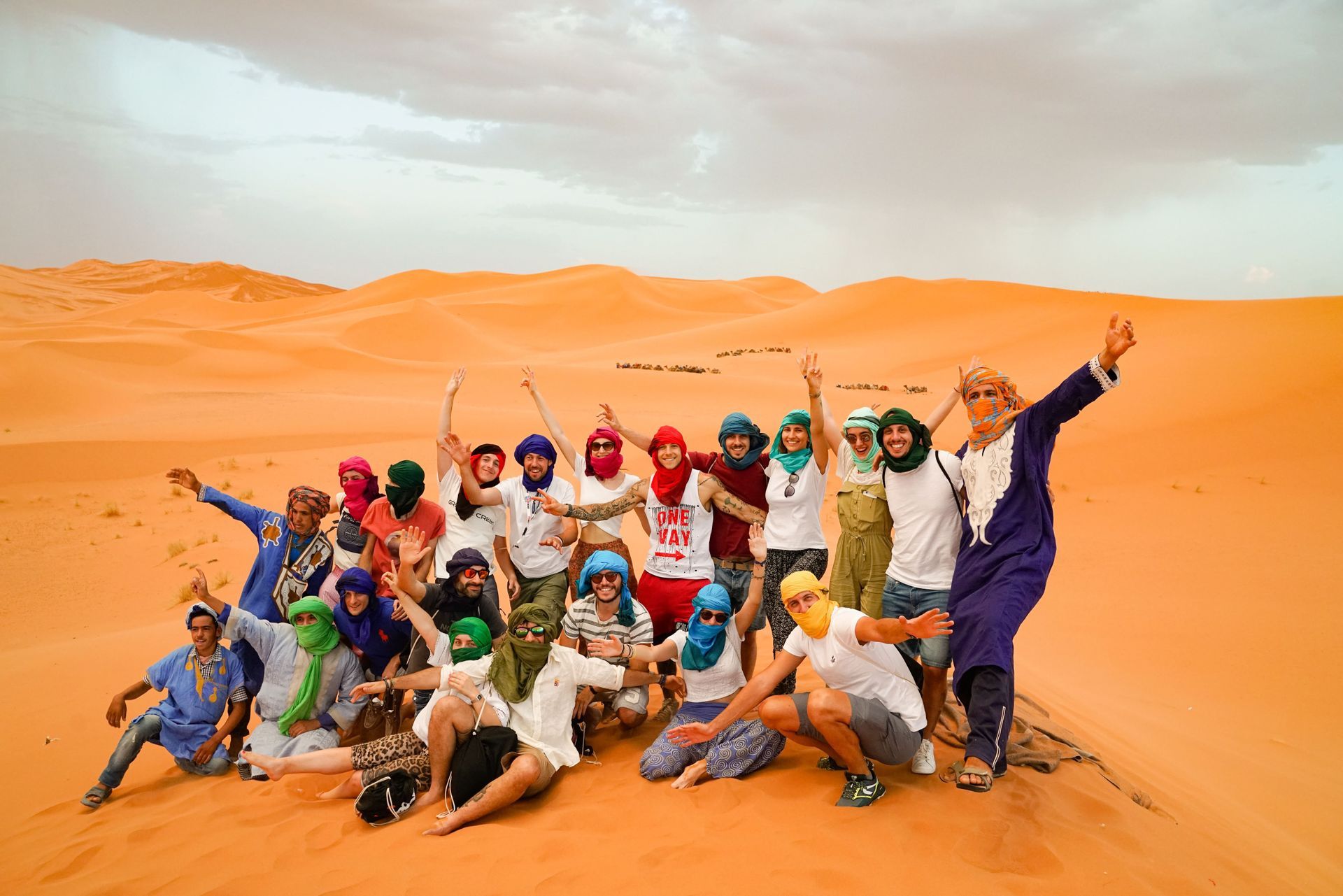 A large WeRoad group trip wearing colorful headscarves and turbans poses for a group photo on a sand dune in the desert.