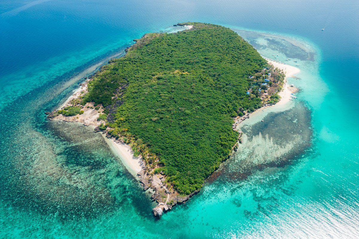 Vista aérea de una isla tropical cubierta de bosque verde, rodeada de agua turquesa y arrecifes de coral.