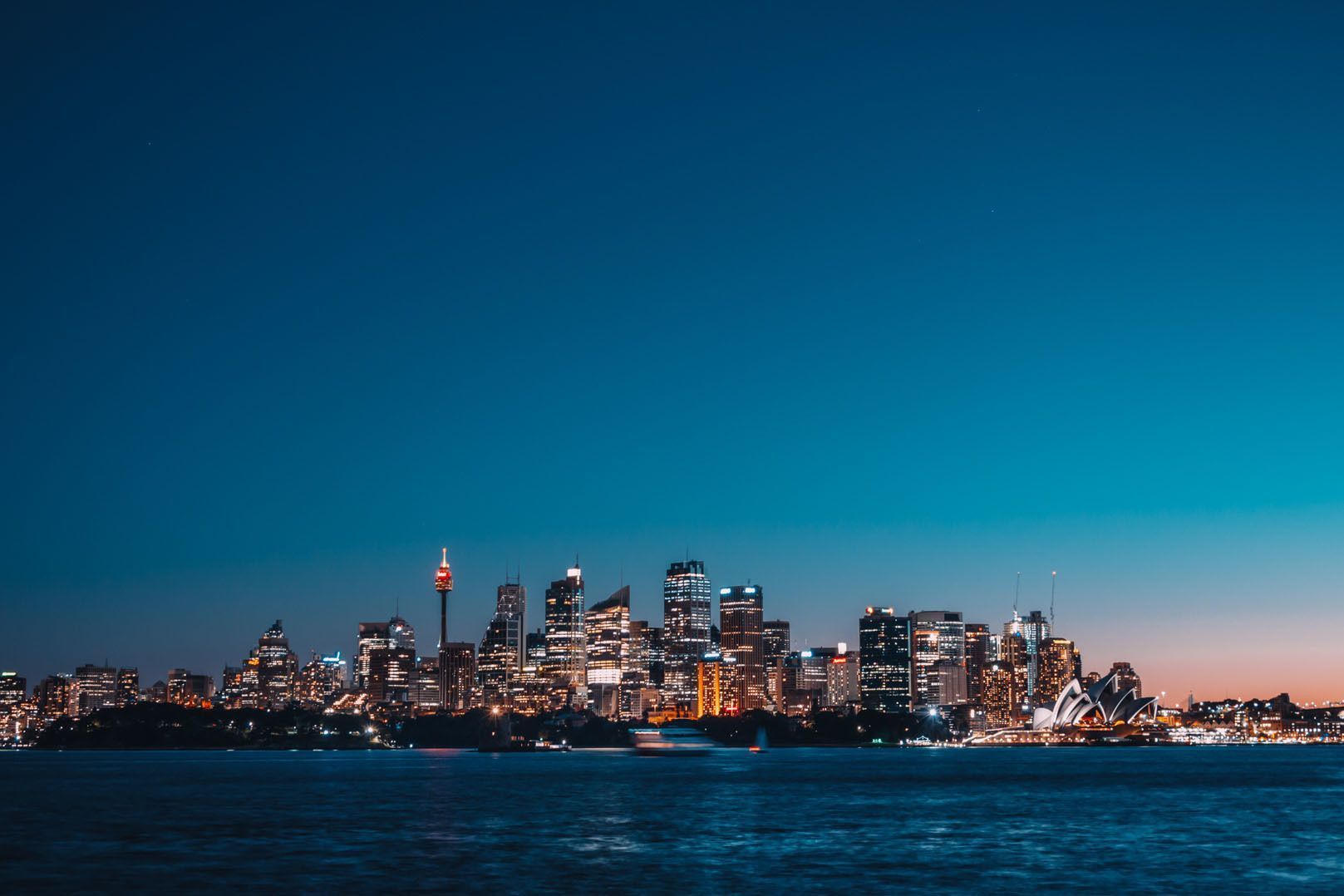 An illuminated city skyline with skyscrapers and modern buildings viewed from across a body of water at dusk.