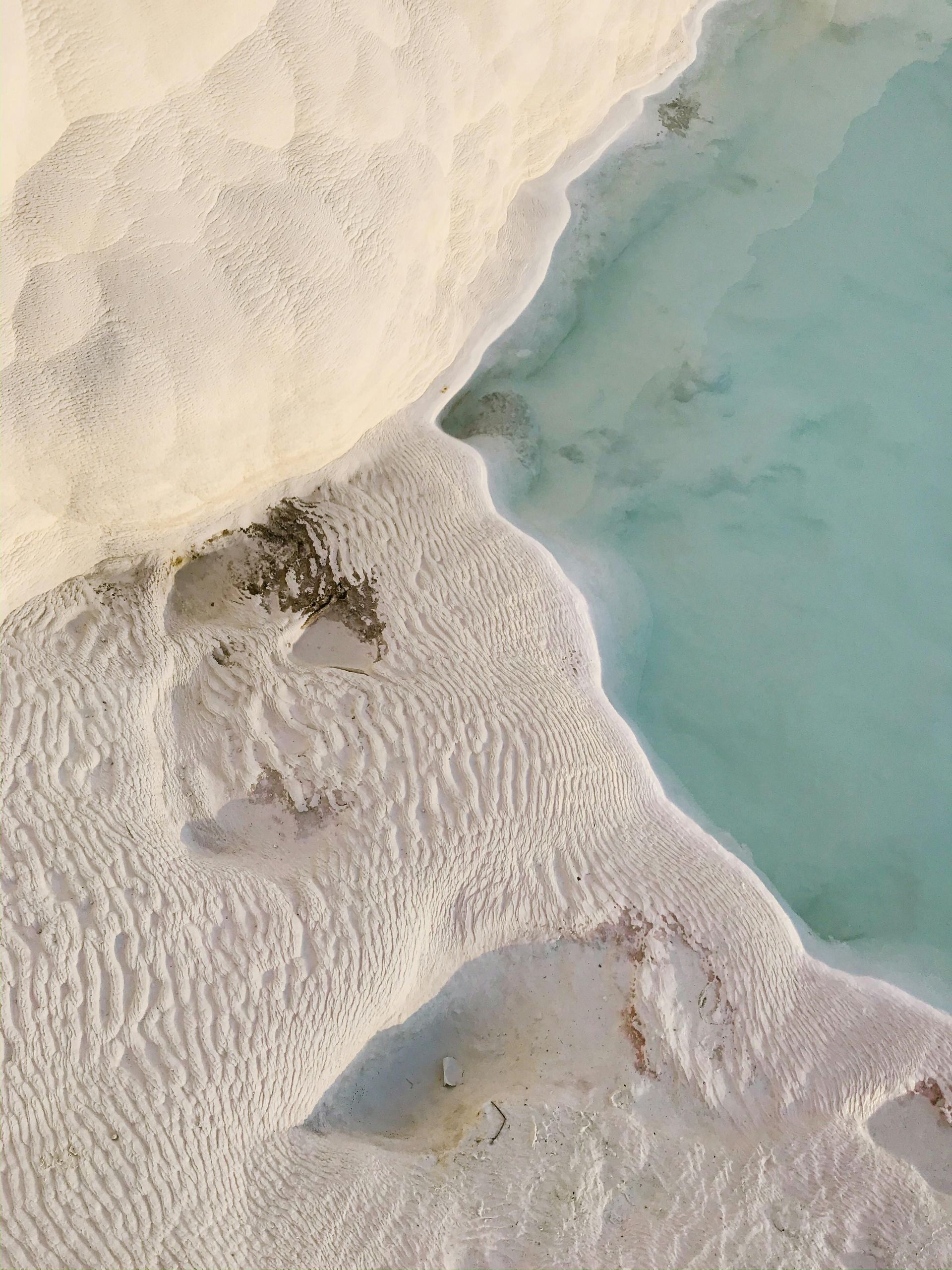 Una vista aerea di depositi minerali bianchi e testurizzati che formano terrazze accanto a uno specchio d'acqua calmo e turchese.