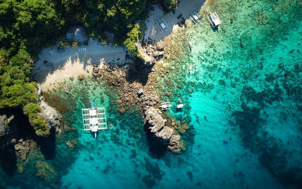 Vista aérea cenital de una cala tropical con barcos sobre agua turquesa, una playa de arena y una costa rocosa con árboles verdes.