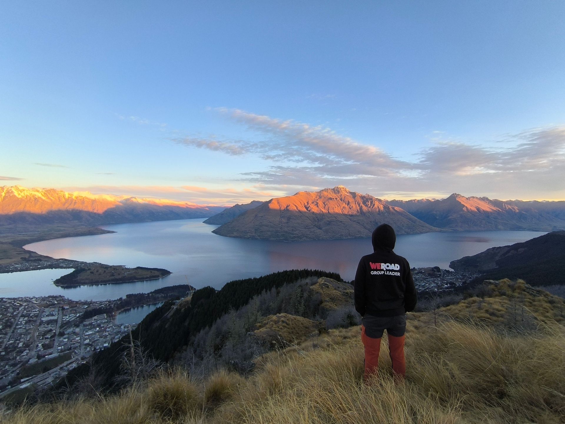 Une personne portant un sweat à capuche noir WeRoad Group Leader se tient sur une colline surplombant un vaste lac entouré de montagnes au lever du soleil.