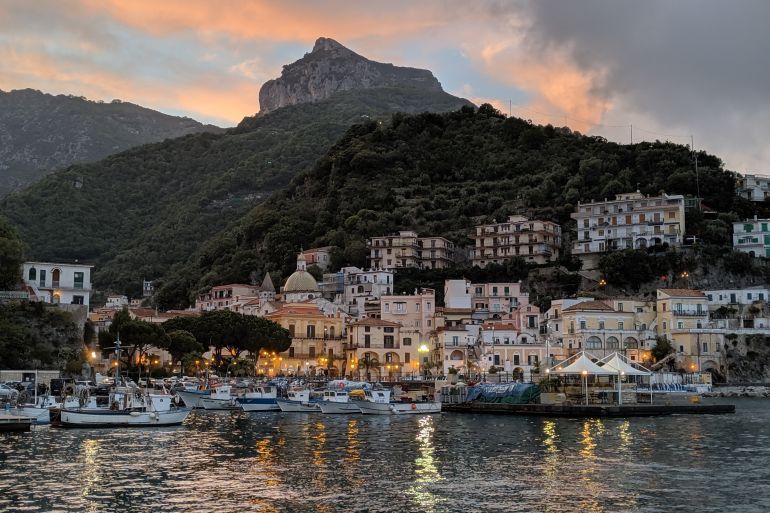 Una città portuale ai piedi di una montagna boscosa, con le luci degli edifici e delle barche che si riflettono sull'acqua al tramonto.