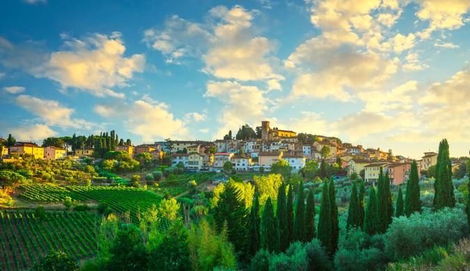 Un borgo illuminato dal sole su una collina circondata da verdi vigneti e cipressi sotto un cielo azzurro con nuvole sparse.