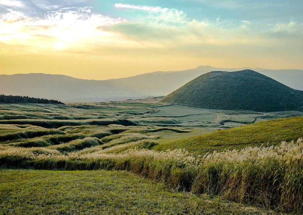 Un paesaggio di dolci colline verdi e un grande monte conico coperto di erba piumosa sotto un cielo velato e colorato.